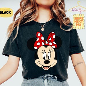 May include: Black t-shirt featuring a Minnie Mouse graphic. The design includes Minnie's face with a red bow with white polka dots. The word "BLACK" is in a yellow oval. The shirt is a dark color.