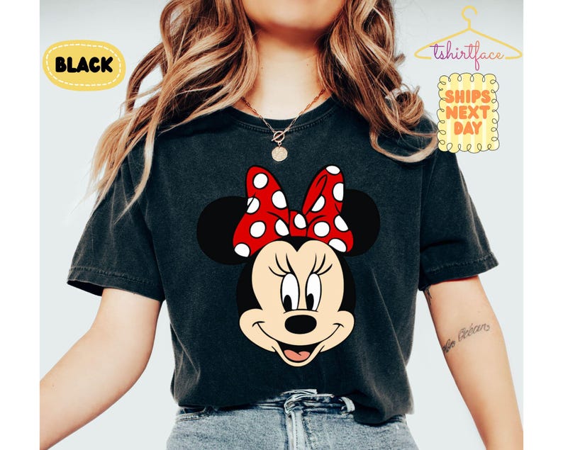 May include: Black t-shirt featuring a Minnie Mouse graphic. The design includes Minnie's face with a red bow with white polka dots. The word "BLACK" is in a yellow oval. The shirt is a dark color.