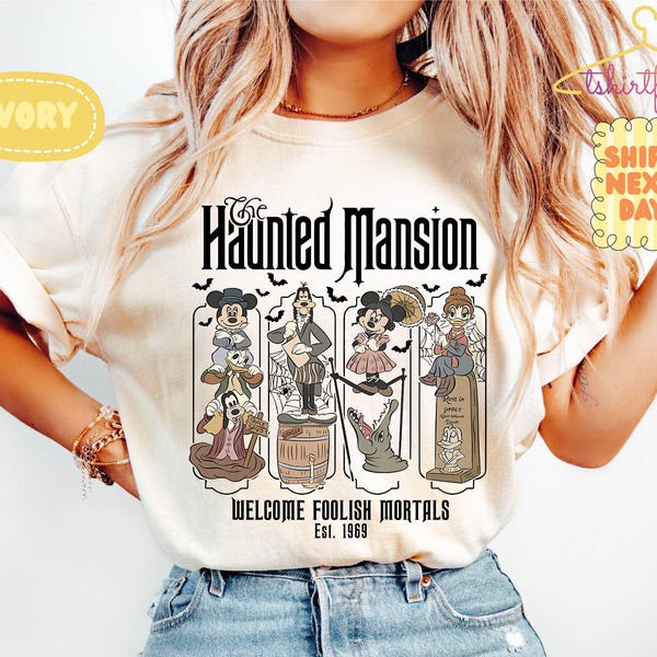 May include: An ivory t-shirt featuring a graphic print of The Haunted Mansion with Disney characters including Mickey, Goofy, and Minnie. The text reads The Haunted Mansion, Welcome Foolish Mortals, and Est. 1969, surrounded by bats and spiderwebs.