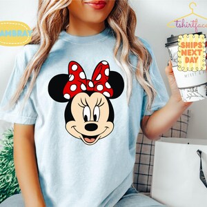 May include: Light blue t-shirt featuring a Minnie Mouse graphic. The graphic shows Minnie's face with a red bow with white polka dots. The shirt has the text "Chambray" and "tshirtface" on it.