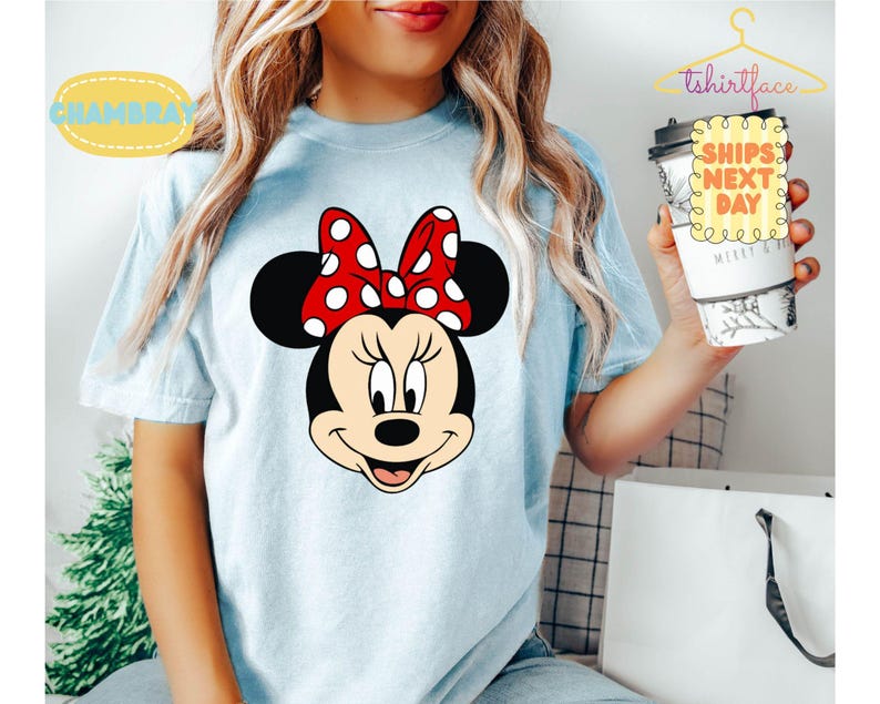 May include: Light blue t-shirt featuring a Minnie Mouse graphic. The graphic shows Minnie's face with a red bow with white polka dots. The shirt has the text "Chambray" and "tshirtface" on it.