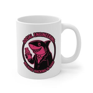 Puede incluir: Taza de cerámica blanca con un tiburón de dibujos animados con traje sosteniendo una unidad USB. El diseño incluye el texto "SOCIAL ENGINEERING" y "BECAUSE HACKING IS HARD" en un círculo rosa.
