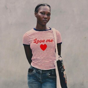 Può includere: Una persona indossa una maglietta a righe rosse e bianche con le parole "Love me" e un cuore rosso. La persona indossa anche jeans blu e porta una borsa tote con un disegno floreale.