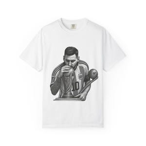 Peut inclure: Un t-shirt blanc à manches courtes avec une illustration graphique en noir et blanc représentant un joueur de football portant un maillot numéro 10, embrassant un trophée, avec un ballon de football placé sur le côté droit du personnage.