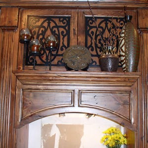 May include: A wooden cabinet with decorative ironwork panels and a shelf displaying various home decor items. The shelf holds glass candle holders, a decorative plate, a vase with branches, and a tall patterned vase. A vase of yellow flowers sits below.