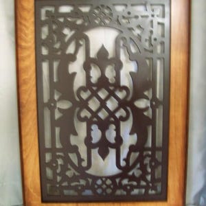 May include: A rectangular decorative wall panel with a dark brown, ornate metal overlay set within a light brown wooden frame. The metal design features a symmetrical pattern of swirling lines and floral elements.