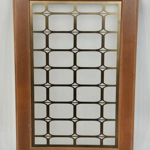 May include: A wooden cabinet door with a brass-coloured decorative insert. The insert has a grid pattern of rectangular and diamond shapes. The wood frame is a medium brown, and the insert is a contrasting gold colour.