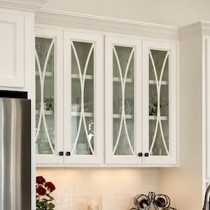 May include: White kitchen cabinets with glass doors featuring a curved mullion design and small black knobs. The interior shelves hold glassware and plates. Below, a black metal wine rack, a candle in a gold holder, and a potted red flower.