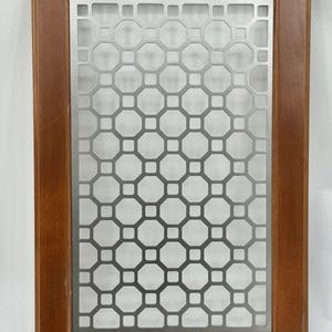 May include: A rectangular wooden cabinet door with a decorative metal insert. The wood frame is a medium brown colour. The metal insert features a repeating geometric pattern of octagons and squares, creating a visually appealing design.