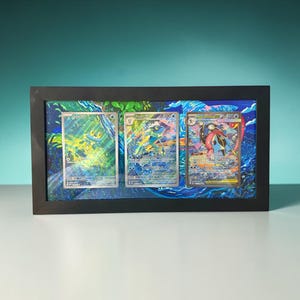 May include: A rectangular black frame containing three collectible trading cards arranged horizontally. The cards feature blue and green creature illustrations with holographic details, set against a vibrant, artistic background of swirling blue and green patterns.