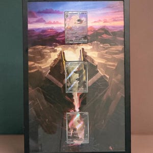 May include: A black rectangular frame containing three vertically aligned trading cards featuring Charizard, Charmeleon, and Charmander. The background art depicts a sunset over a rocky landscape with a glowing fissure running down the center.