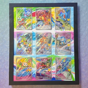 May include: A black rectangular frame containing nine holographic Pokémon trading cards arranged in a three-by-three grid. The cards feature various colorful animated characters including Bulbasaur, Charmander, Squirtle, Turtwig, Chimchar, Piplup, Rowlet, Litten, and Popplio, all set against a vibrant, multi-colored graphic background.