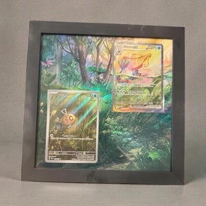 May include: A black square frame containing two Pokémon trading cards, Feebas and Milotic, displayed over a vibrant, detailed forest background illustration. The cards feature holographic elements and text, with Feebas on the left and Milotic on the right, creating a layered, three-dimensional effect within the display.