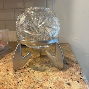 May include: A clear, round crystal bowl with a starburst pattern sits in a silver metal stand. The stand has three legs with a unique, stylized design. The bowl is likely for decorative purposes.