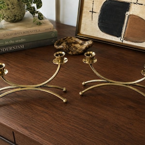 May include: A pair of polished brass candle holders, each featuring a curved, arching base with four rounded feet and a single central candle cup. The metallic gold-toned finish reflects light, and the design creates a symmetrical, balanced appearance for holding taper candles on a flat surface.