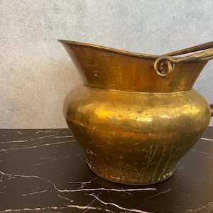 Pode incluir: Um jarro de latão vintage com corpo arredondado e borda alargada. O jarro tem uma alça e um bico. O latão tem um tom dourado quente com alguns sinais de idade e desgaste. O jarro é provavelmente usado para fins decorativos.