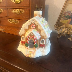May include: A ceramic gingerbread house figurine featuring white icing details, a candy cane arched doorway, red and green gumdrop accents, and a roof decorated with colorful pastel candy circles and a central white chimney stack.