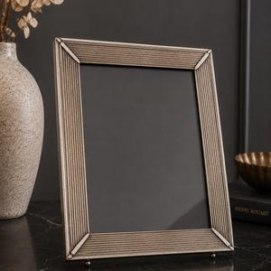 May include: A rectangular picture frame with a metallic silver finish and a textured, ribbed surface pattern. The frame features mitered corners and stands on small, rounded feet, surrounding a dark, empty central display area for a photograph.