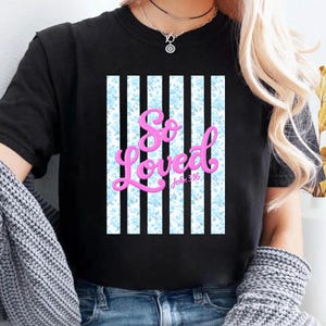 Puede incluir: Camiseta negra con un diseño gráfico. El diseño presenta las palabras "So Loved" en escritura rosa, con "John 3:16" debajo. El fondo es un patrón de rayas verticales con acentos florales azules.