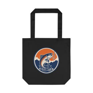 May include: Black tote bag with a circular graphic featuring a jumping fish, a fishing hook, and a mountain range against an orange and blue background. The bag has two black handles.