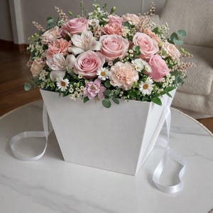 Puede incluir: Un arreglo floral en una caja cuadrada blanca. El ramo presenta rosas rosas, claveles y margaritas, con vegetación. Cintas blancas caen de la caja, colocada sobre una mesa con tapa de mármol.