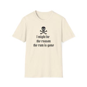 May include: Cream-colored t-shirt featuring a black skull and crossbones graphic above the text "I might be the reason the rum is gone." The shirt has a crew neck and short sleeves. The fabric has a speckled texture.