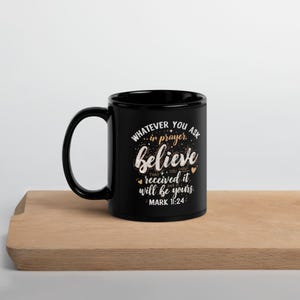 May include: A black ceramic mug with a white inspirational quote. The text reads "Whatever you ask in prayer, believe that you have received it and it will be yours. Mark 11:24". The mug is on a wooden surface.