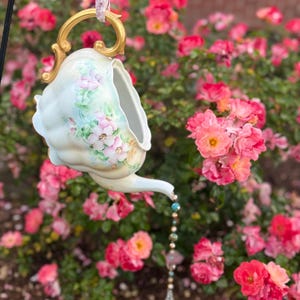 May include: A vintage-style, hand-painted ceramic teapot bird feeder with a gold handle and spout. The teapot is decorated with pink floral designs and hangs from a floral ribbon. A beaded charm hangs from the spout. The background features a vibrant rose bush.