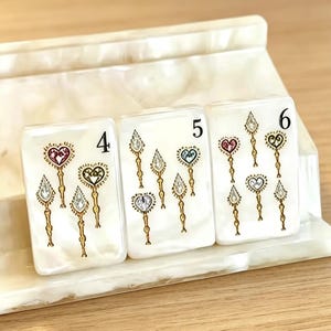 Puede incluir: Tres fichas de juego rectangulares blancas con diseños ornamentados de oro y joyas. Cada ficha presenta un número: 4, 5 y 6. Los diseños incluyen formas de corazón y lágrima con detalles de joyas. Las fichas están sobre una superficie blanca texturizada.