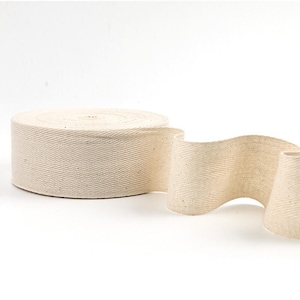 May include: A roll of natural cotton webbing with a herringbone weave. The webbing is unrolled, displaying its texture and flexibility. Suitable for various craft and sewing applications. The roll is in a neutral colour.