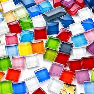 May include: A collection of small, square glass mosaic tiles in a vibrant array of colours including red, blue, green, yellow, orange, pink, and clear. The tiles have a smooth, glossy finish and are scattered across the frame in a random arrangement.