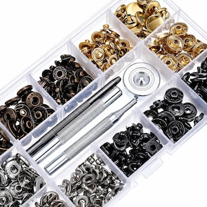 May include: A clear plastic organiser containing an assortment of metal snap fasteners in gold, bronze, and black. Includes silver metal tools for setting the snaps. The fasteners are in various shapes and sizes, suitable for sewing and craft projects.