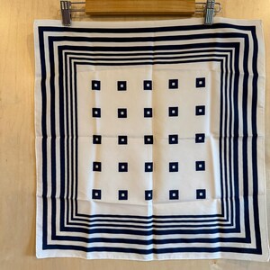 May include: White square scarf with a navy blue border and navy blue square accents. The scarf is hanging on a wooden hanger. The scarf has a classic, geometric design.