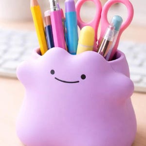 May include: A light purple desk organizer shaped like a blob with a simple face featuring two black dots for eyes and a curved line for a mouth. The container holds various office supplies including a yellow pencil, pens, and pink scissors.