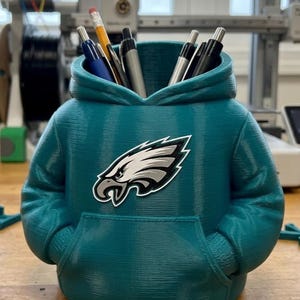 Può includere: Un organizer da scrivania color verde acqua stampato in 3D a forma di felpa con cappuccio. La parte anteriore presenta il logo dei Philadelphia Eagles in bianco, nero e argento. Il design include una tasca a marsupio e un'apertura per le penne.