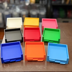 May include: A collection of nine square, open-top trays arranged in three rows. The trays are available in various solid colors including white, yellow, pink, black, red, green, dark blue, orange, and light blue, each featuring a notched front edge.
