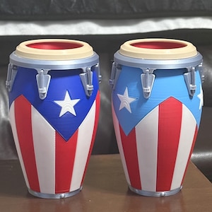 May include: Two conga drums featuring a Puerto Rican flag design with vertical red and white stripes, a blue triangle, and a white star. The drums have silver-colored hardware, beige rims, and red interiors, standing side by side on a flat surface.