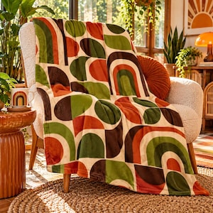 May include: A soft throw blanket featuring a repeating geometric pattern of semicircles and arches in shades of burnt orange, olive green, and dark brown on a cream background, draped over a textured white armchair in a bright indoor setting.