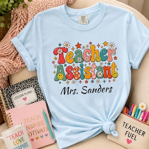 Puede incluir: Una camiseta azul claro con el texto Teacher Assistant en una fuente colorida de estilo retro, decorada con pequeñas flores, estrellas y caras sonrientes. Debajo del texto principal, el nombre Mrs. Sanders está impreso en letra cursiva negra.
