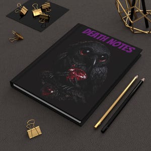 Peut inclure: Journal noir avec le texte "DEATH NOTES" en violet. La couverture présente une illustration sombre d'un hibou aux yeux rouges et un bijou rouge. Des crayons dorés et noirs et des pinces à reliure dorées sont également visibles.