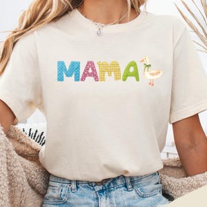 Könnte beinhalten: Cremefarbenes T-Shirt mit dem Wort "MAMA" in bunten, gemusterten Buchstaben und einer kleinen Entengrafik. Das Shirt ist kurzärmelig und hat eine lockere Passform. Das Wort "Natural" ist auf einem cremefarbenen Hintergrund gedruckt.