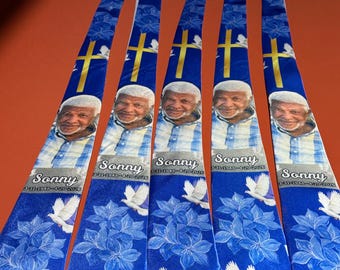 May include: A collection of blue neckties with a personalized design. Each tie features a portrait, the name "Sonny", and dates. The ties also display a gold cross, white doves, and floral patterns. The ties are hanging on a white hanger.