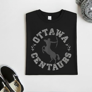 May include: A black t-shirt featuring a circular graphic with the text OTTAWA CENTAURS in a white patterned font. In the center, a solid gray silhouette of a centaur archer aiming a bow is displayed. The design includes small leaf accents.
