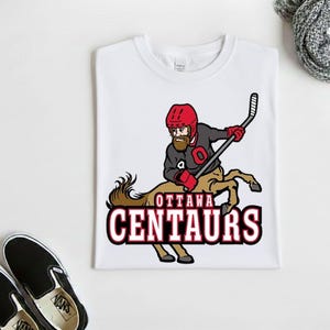 May include: A white t-shirt featuring a graphic of a centaur hockey player wearing a red helmet and black jersey with a red letter O. The centaur holds a hockey stick, and the text OTTAWA CENTAURS is printed in red and black below the figure.