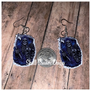 May include: A pair of dangle earrings featuring a grim reaper design with a scythe and skulls, in shades of blue and black. The earrings have silver-colored hooks. The text "Skull Crusher Designs" is visible.