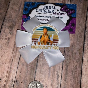 May include: A white satin bow with a circular image of a man with the text "NOW THAT'S WHAT I CALL HIGH QUALITY H2O". The bow is on a wooden surface with a quarter dollar coin.