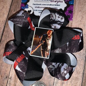 May include: A large, handmade hair bow with a black and white patterned ribbon, featuring a small image of a character holding a baseball bat. The bow has red accents and the words "The Walking Dead" printed on it. A quarter is next to the bow.