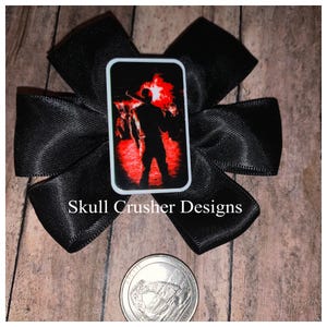 May include: Black satin hair bow with a rectangular image of a person with a baseball bat in front of a red background. The text "Skull Crusher Designs" is printed on the bow.