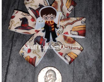 May include: A brown, white, and red bow with a Harry Potter character in the center. The bow is made of ribbon with a Harry Potter themed print. The character is wearing a black robe and glasses. The bow is about the size of a quarter.