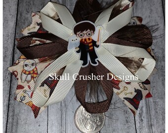 May include: A brown, white, and gold hair bow with a Harry Potter character charm. The charm features a cartoon image of Harry Potter holding a wand and a white owl. The bow is decorated with a brown and white striped ribbon and a brown and white patterned ribbon. The text "Skull Crusher Designs" is visible on the bow.